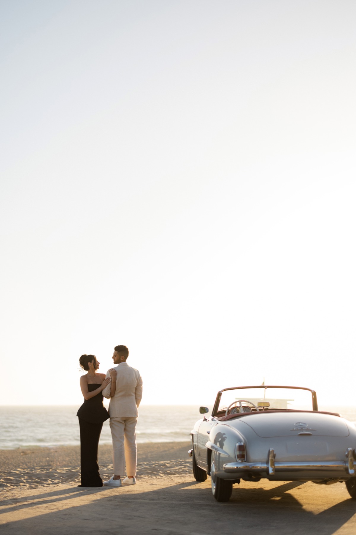 Couple standing beside the Mercedes 190 SL on a Malibu beach at sunset