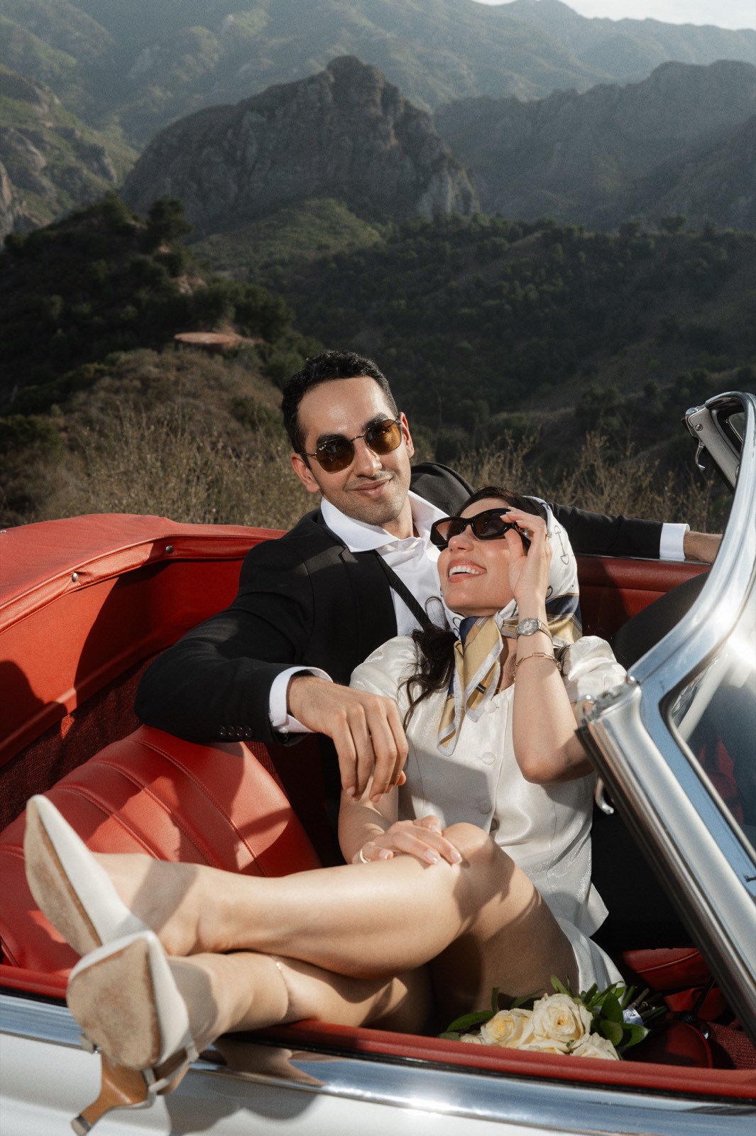 Couple in Riviera style with silk headscarf lounging in the red leather seats of the 190 SL